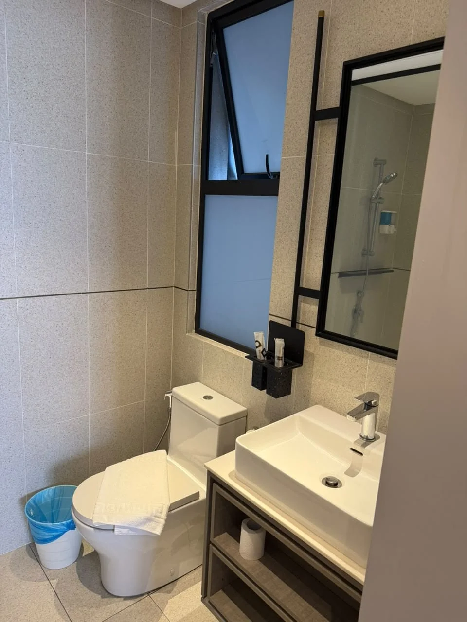 Bathroom features: white ceramic toilet, single-sink vanity with wood cabinet, chrome basin mixer faucet, framed mirror, shower with glass enclosure, tile cladding on walls, light-colored tile flooring, window for natural light, blue waste bin; style: modern.